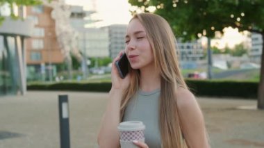 Uzun sarı saçlı mutlu genç bir kadın parkta erkek arkadaşıyla telefonda konuşuyor. Bayan neşeyle gülüyor, dışarıda kahve içiyor.