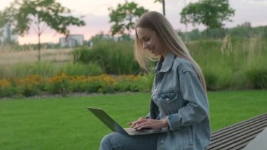 Kot ceketli sarışın genç kadın bilgisayardaki arkadaşına mutlu bir şekilde gülümsüyor. Neşeli bayan internette dizüstü bilgisayarda sörf yapıyor park bankında oturuyor.