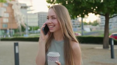 Elinde kahve fincanı olan neşeli genç bir kadın telefonda erkek arkadaşıyla konuşurken gülüyor. Bayan işten sonra şehir parkında kahve içerek vakit geçiriyor.