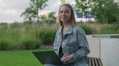 Kucağında dizüstü bilgisayarı olan genç sarışın kadın akşam vakti bankta oturan kameraya bakıyor. Kot ceketli kadın işten sonra parkta dinleniyor.