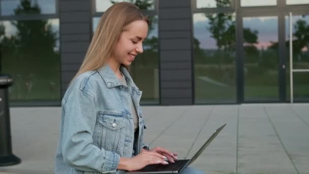 穿着斜纹棉布夹克的年轻女性微笑着在城市街道上自由工作 膝上型笔记本电脑 放松快乐的女士在工作休息的时候给朋友发短信 — 图库视频影像
