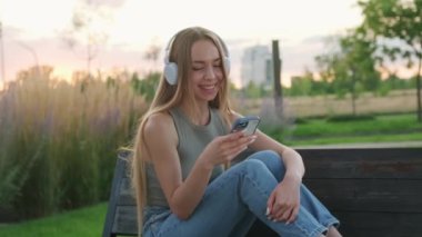 Kulaklıklı neşeli bayan yazın bankta bacaklarına sarılıyor. Genç bir kadın akşam saatlerinde arkadaşlarıyla mesajlaşırken müzik dinler.
