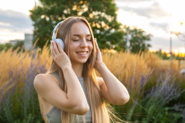 Neşeli genç kadın gün batımında parkta otururken müzik dinlemekten ve şarkı söylemekten hoşlanır. Kulaklıklı mutlu bayan doğada zaman geçiriyor.