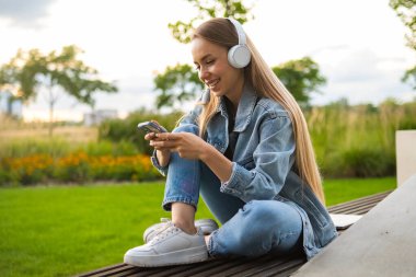 Akıllı telefonu olan genç bir kadın parktaki bankta oturuyor. Kot ceketli kadın işten sonra parkta dinleniyor.