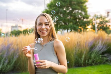 Şehir bahçesinde sabun köpüğü üfleyen neşeli genç bir kadın.