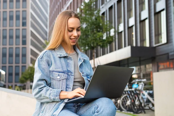 Stock image Young woman in denim jacket smiles working freelance on city street with laptop on lap. Relaxed happy lady texts friends at work break