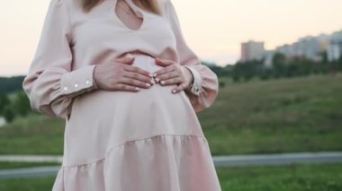 Müstakbel anne çocuk beklentisiyle karnını nazikçe okşar. Kadın alacakaranlıkta şehir binalarına karşı tepelik çimlerde duruyor.