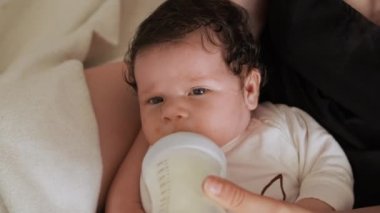 Anne küçük çocuğa plastik şişeden süt içmesi için verir. Bebek anne kollarında yatarken vitaminlerle dolu besleyici içecekler tüketir.