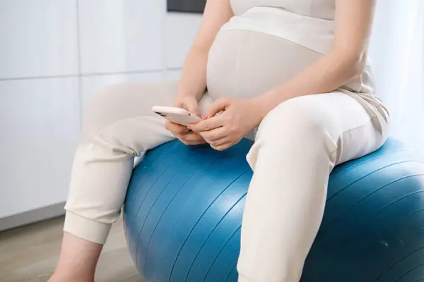 Pregnant woman moves slowly gently rocking body while sitting on rubber ball. Future mother performs gymnastic exercises checking phone during movements