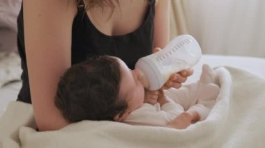 Tişörtlü anne yeni doğmuş bebeği plastik şişeden süt ile besler. Çocuk anne kollarında uzanıp vitaminlerle beslenen yiyecekler yiyor.