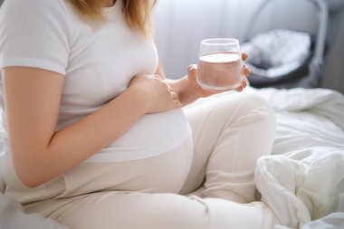 Hamile kadın yatakta oturuyor, nazikçe karnını kucaklıyor ve elinde bir bardak su tutuyor..