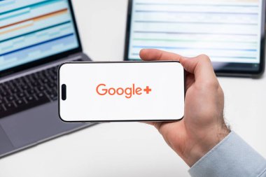 Google Plus logosu, arka planda grafiksel diyagramlı tablet ve dizüstü bilgisayarlı akıllı telefonun ekranında, Aralık 2023, Prag, Çek Cumhuriyeti.
