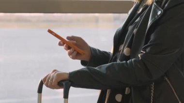 Siyah ceketli ve benekli elbiseli bir kadın havaalanında elinde bavulla cep telefonunu kaydırıyor.. 