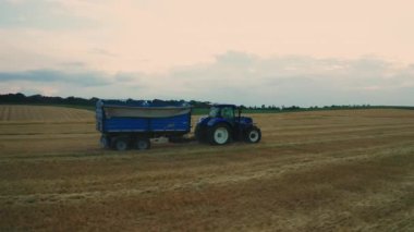 Tramvaylı traktör, ekili tarımsal alan manzarası boyunca ilerliyor. Kamyon nakliyecileri yaz akşamları tarım çiftliğinde mahsul topluyor.