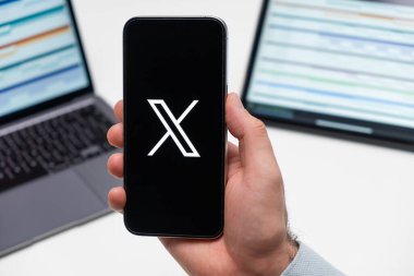 Platform X sosyal medya uygulamasının logosu erkek eli ekranında, dizüstü bilgisayar ve tablet arka planda, Aralık 2023, Prag, Çek Cumhuriyeti