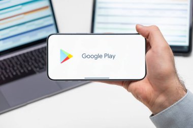 Google Play uygulamasının logosu bilgisayarın ve tabletin önünde bulunan adamın cep telefonu ekranında, Aralık 2023, Prag, Çek Cumhuriyeti
