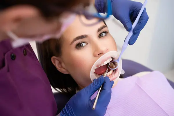 歯科医は 女性患者の歯をチェックするために歯科用具と鏡を使用しています ストック画像