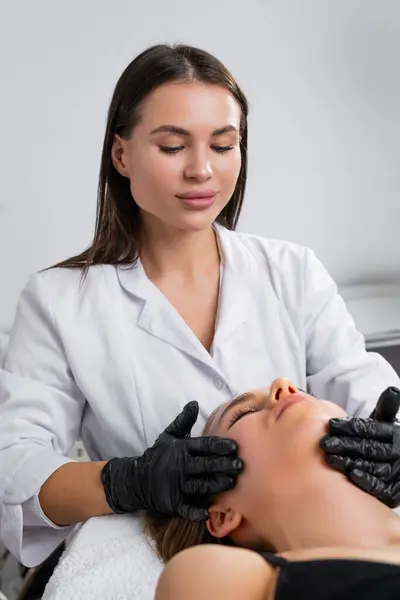 美容院で若い女性化粧品師が若い女性を嘘をつくために顔の治療をしている ロイヤリティフリーのストック画像