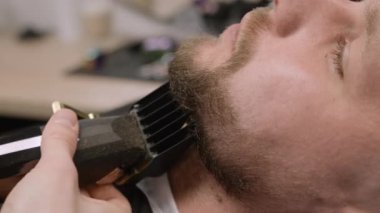 Stilist, berber dükkanında çalışan genç adamın sakalını kesiyor. Profesyonel berber kuaförde erkek müşterinin yüz kıllarını kesiyor.