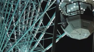 Rotating Ferris wheel with hanging cabins and sparkling LED lights at night. Popular attraction for fair guests against black sky. Amusement park