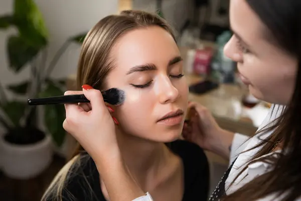 Makeup artist lightly applies highlighter with special brush on client face.