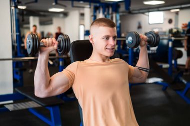 Genç vücut geliştirici spor salonundaki bir bankta otururken dambılları kaldırır.