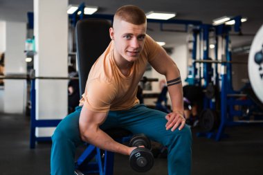 Vücut geliştirici, spor salonundaki bir bankta otururken halterle biseps kıvırır.