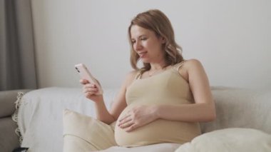 Mutlu hamile kadın akıllı telefondan sosyal medyada göbeğini okşuyor. Genç bayan koltukta oturarak işe yarar annelik ipuçları arıyor.