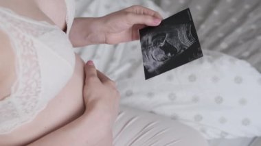 Hamile kadın karnını nazikçe okşarken yatak odasında ultrason görüntüsü tutuyor. Müstakbel anne yatakta oturan bebeğe dokunuyor.