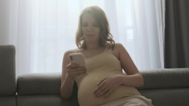 Hamile kadın akıllı telefondan kitap okurken karnını nazikçe yakıyor. Sevimli gülüşü olan mutlu genç kadın, aktif bebeği, nazik dokunuşlarla sakinleştirir.