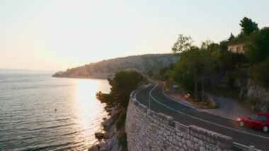 İki yolcu aracı, alacakaranlıkta deniz koyuna kurulmuş kıyı yolunda ilerliyor. Güneş ışınları dağlık alana karşı suyu yansıtır.