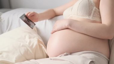 Hamile kadın doktor muayenesinden sonra ultrason görüntüsüyle kanepede oturuyor. Rahatlamış genç dişi nazikçe koca göbeğini okşar ve anın tadını çıkarır.