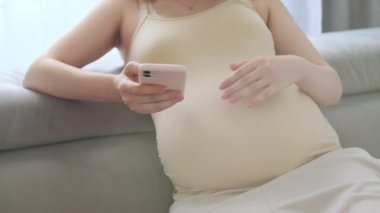 Hamile kadın akıllı telefonu tutarak karnını nazikçe okşuyor. Genç bir kadın internette işe yarar annelik ipuçları arıyor koltukta oturuyor.