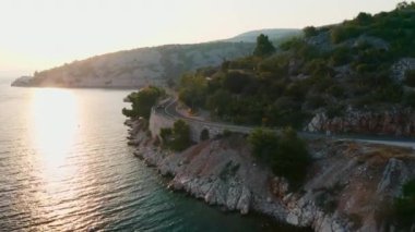 Yolcu vagonu, deniz kenarındaki çift şeritli otobandan geçiyor. Güneş ışığını yansıtıyor. Yamacın yamacında suyun yanında, arkaplan manzaralı dağlar var.