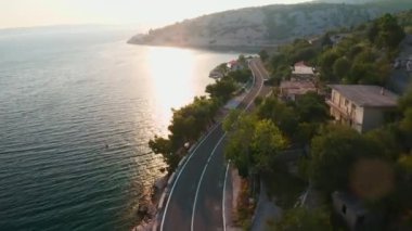 Şafakta sahil yolu boyunca giden bir cip. Dağ tepelerindeki köy evleri ve seyrek ormanlar kıyı havası manzarası yakınındaki yol çevresini sarar.