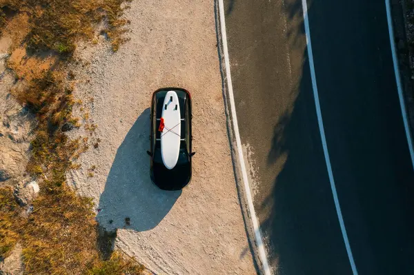 stock image Black car with a paddleboard securely fastened on its roof, parked neatly on the shoulder of a winding coastal road. 