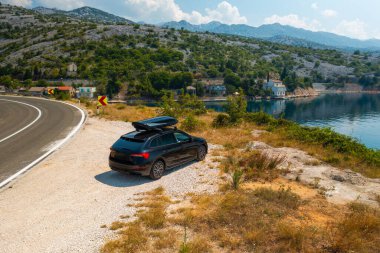 Uçurumun kenarındaki dolambaçlı bir yola park etmiş siyah bir araba. Parlak güneş ışığı altında masmavi deniz parlıyor.