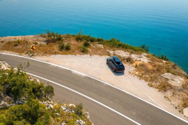 Tepe kenarındaki bir yolun kenarına park edilmiş, açık mavi gökyüzünü yansıtan masmavi bir deniz olan bir araba.. 