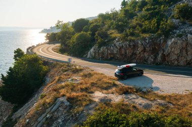 Çatıya bantlı siyah bir araba, uçurumun kenarına park edilmiş, aşağısı gök mavisi.