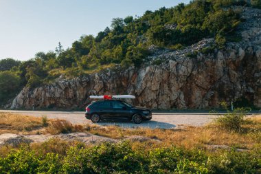 Çatısına SUP bağlanmış siyah bir araba, dolambaçlı bir kır yolunun kenarında duruyor.