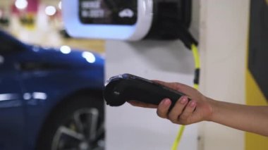 Kadın, istasyonda elektrikli araç şarj servisi için akıllı telefonla ödeme yapıyor. Müşteri para transfer ederken kasiyer POS terminalini tutar