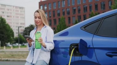 Şehir istasyonunda elektrikli arabanın yanında akıllı telefonu ve cam su şişesi olan mutlu bir kadın. Sarışın bayan sorumluluk sahibi çevre dostu yaşam tarzını seviyor.