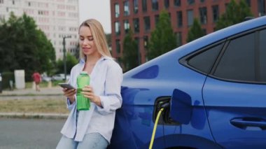 Sarışın kadın, şehir istasyonunda elektrikli arabaya yaslanarak gülümsüyor. Kadın sürücü EV 'nin yanında yeniden kullanılabilir su şişesi ve akıllı telefon tutuyor.