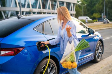 Woman charging her electric car while carrying groceries near a modern shopping center.  clipart