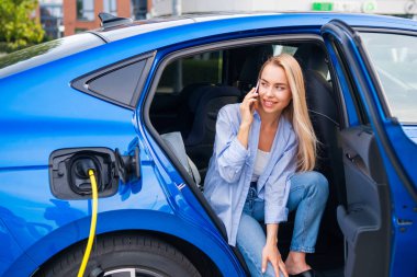 Kadın elektrikli arabasında oturuyor, şarj ediyor ve dışarıda akıllı telefonuyla konuşuyor..