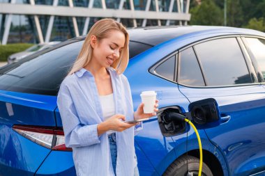 Kadın akü seviyesini kontrol etmek için akıllı telefonunu kullanıyor ve elektrikli arabası şarj olurken kahve içiyor..