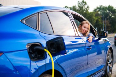 Elektrikli araç caddedeki bir istasyonda şarj olurken sarışın kadın pencereden sarkıyor.