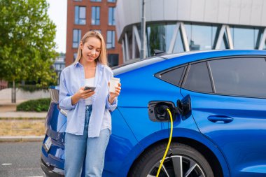 Genç bir kadın kahvesini içerken elektrikli arabasını şarj ediyor ve akıllı telefonunu sokakta ödeme olarak kullanıyor.
