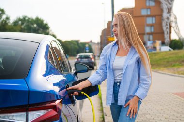 Uzun saçlı sarışın bir kadın, elektrikli arabasını cadde kenarındaki bir şarj aletine bağlıyor.