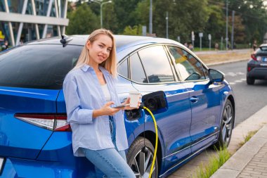 Sarışın kadın, günlük kıyafetler giymiş, elektrikli arabanın yanında duruyor, kahvesini yudumluyor ve akıllı telefonunu kontrol ediyor.